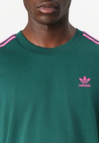Zielona bawełniana koszulka z krótkim rękawem, z różowymi paskami Adidas na ramionach oraz małym różowym logo na piersi. Gładka faktura.
