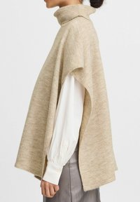Beige turtleneck poncho met een losse pasvorm en textuur brei. De stof valt elegant over de schouders, gecombineerd met een wit shirt eronder.