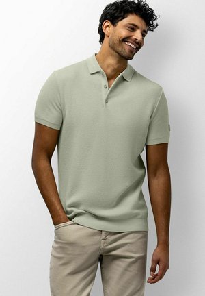 Man smiling wearing light green short-sleeve polo shirt and beige pants, standing with one hand in pocket against white background.