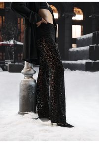 Femme portant un pantalon en dentelle noire et des talons hauts, debout sur la neige à côté d'un lampadaire avec un bâtiment en arrière-plan.