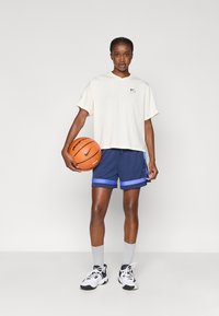 Λευκή αθλητική μπλούζα με κοντά μανίκια, μπλε σορτς μπάσκετ με μπλε λεπτομέρειες, γκρίζες κάλτσες, μαύρα και άσπρα αθλητικά παπούτσια, κρατώντας μια πορτοκαλί μπάλα μπάσκετ.