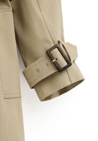 Beige trench coat sleeve with adjustable fabric strap and dark rectangular buckle detail on a white background.