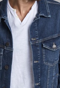 Denim jacket in dark blue with front pockets, button closure, and distressed details, layered over a white v-neck shirt.