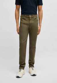 Olive green trousers with a slim fit, five pockets, zip fly, and button closure. Worn with a navy shirt and cream sneakers.