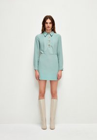 Abito verde menta con maniche lunghe, chiusura con bottoni e colletto alto. Abbinato a stivali beige fino al ginocchio. Texture liscia e vestibilità su misura.