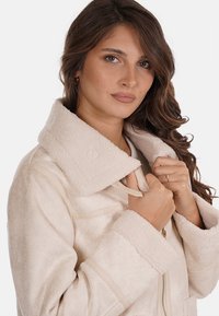 Giacca in ecopelliccia color crema con colletto alto, chiusura con zip e hardware dorato a contrasto. Presenta una consistenza morbida e cuciture dettagliate.