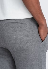 Pantalones grises texturizados con un sutil patrón de espiga, que cuentan con un bolsillo trasero y un corte relajado, llevados con una camisa blanca.