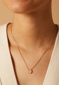 Gouden ketting met een ovaal hangertje met een gladde, lichtgekleurde steen, weergegeven op een halslijn met een geweven trui.