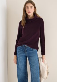 Lila Pullover mit Kapuze, aus leichtem Stoff, kombiniert mit blauen weiten Jeans, hält eine beiger Fleecejacke. Schlichtes, lässiges Design.