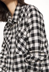 Chemise en flanelle à carreaux noir et blanc avec un col pointu, une patte de boutonnage sur le devant et deux poches poitrine. Texture douce avec les manches retroussées.