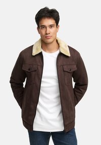 Braune Kunstwildlederjacke mit einem cremefarbenen Lammfellkragen, ausgestattet mit zwei Brusttaschen und einem Frontreißverschluss. Legere Passform, getragen über einem weißen T-Shirt.