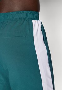 Teal sports shorts med tekstureret overflade, der har en lodret hvid stribe, elastisk talje og en sidelomme med lynlåsdetaile.