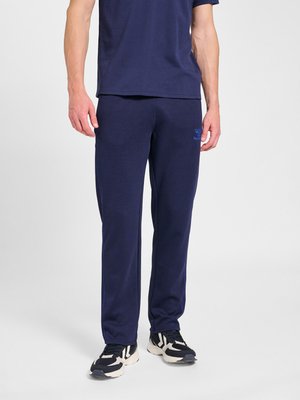 Man draagt marineblauwe sweatpants, bijpassend t-shirt en zwart-witte sneakers, staand tegen een lichte achtergrond.
