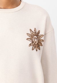 Beige sweatshirt with ribbed neckline and decorative embroidered flower patch. The patch features bronze sequins and a heart shape at its center.