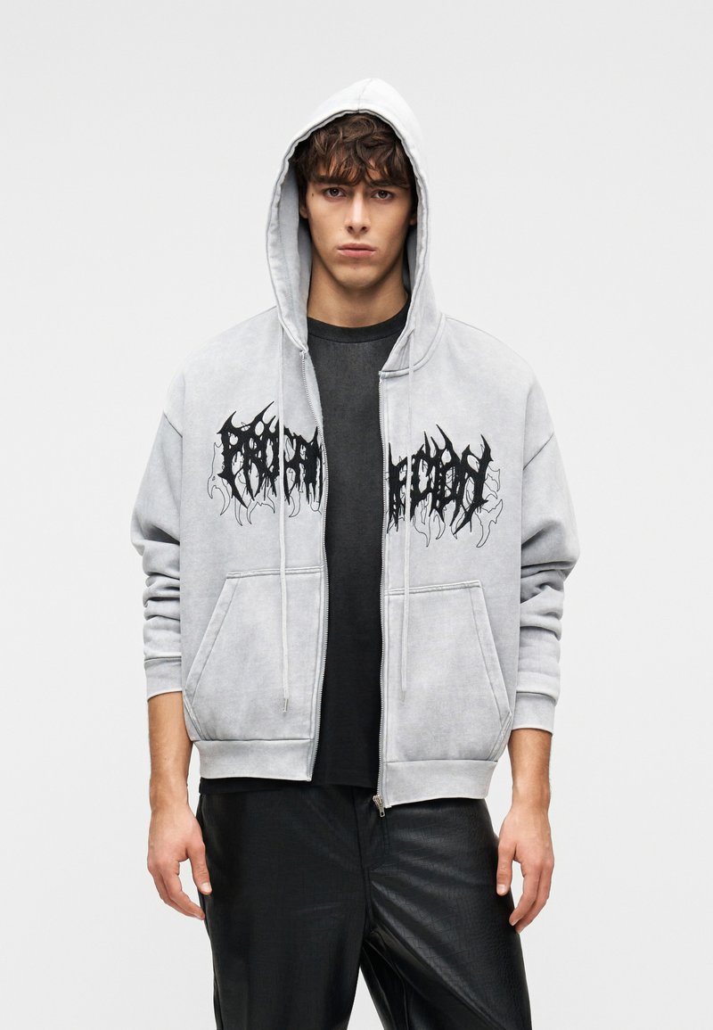 Γκρι φούτερ με φερμουάρ και κουκούλα, με μαύρο γραφικό κείμενο στο μπροστινό μέρος, τσέπες καγκουρό και ριμπ μανσέτες. Φορεμένο πάνω από μαύρο πουκάμισο.