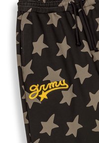 Zwarte trainingsbroek met grote beige sterrenpatronen, een elastische tailleband en gele geborduurde "grmu" tekst met een steraccent.