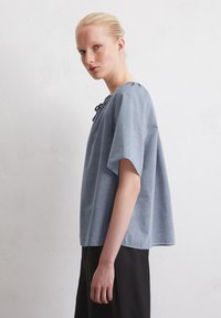 Top azzurro chiaro a maniche corte con una vestibilità morbida, scollatura rotonda e dettaglio con nodo. Realizzato in un tessuto morbido e strutturato, abbinato a una gonna scura.