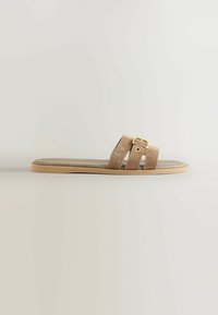 Sandale en daim beige avec deux larges brides, dotée d'une boucle en métal doré. Semelle plate de couleur naturelle, design simple, sans motifs supplémentaires.