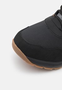 Scarpa sportiva nera caratterizzata da una tomaia testurizzata, suola in gomma con accenti marroni e un design imbottito. Include un'area in mesh flessibile e traspirante.