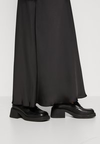 Zwarte chunky hakken met een gladde leren afwerking, met een ronde neus en een decoratieve naaddetail; gecombineerd met een vloeiende zwarte rok.