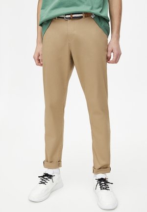 Mann trägt beige Chino-Hosen, an den Knöcheln hochgekrempelt, ein grünes Hemd eingesteckt, einen schwarz-weißen Gürtel, weiße Socken und weiße Sneakers mit schwarzen Schnürsenkeln.
