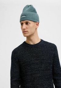 Lyseblå ribbet strikket hue med rund top og broderet logo. Bæres sammen med en tekstureret marineblå crewneck sweater.
