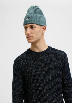Lyseblå ribbet strikket hue med rund top og broderet logo. Bæres sammen med en tekstureret marineblå crewneck sweater.