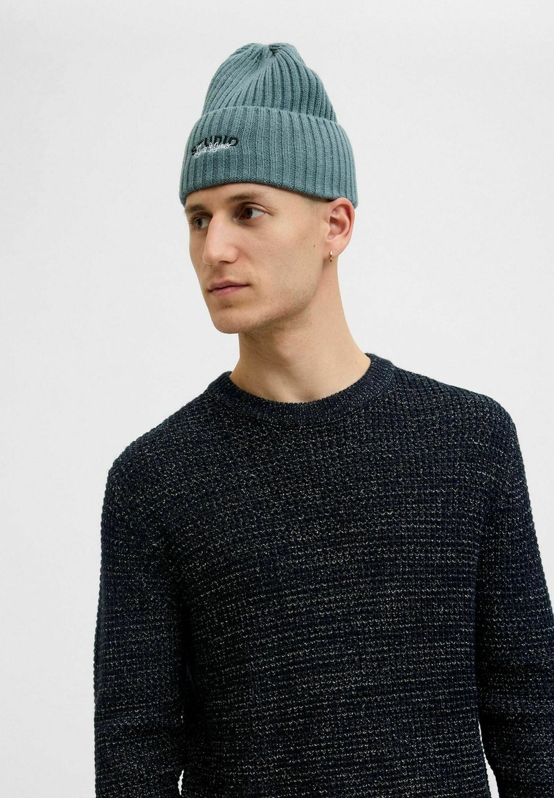 Lyseblå ribbet strikket hue med rund top og broderet logo. Bæres sammen med en tekstureret marineblå crewneck sweater.