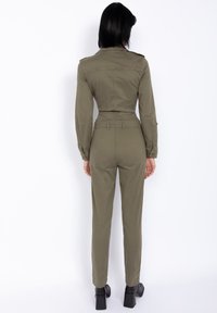 Tuta verde oliva con maniche lunghe, vita aderente e gambe affusolate. Presenta spalline e tasche posteriori, realizzata in tessuto morbido.
