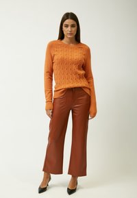Maglione arancione a coste, pantaloni ampi in finta pelle marrone, scarpe con tacco nere. Texture morbide con polsini a costine e una vestibilità rilassata.