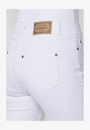 Balti džinsai su siuvinėta užpakaline kišenės iškirpte, metaliniais akcentais ir odiniu ženkliuku juosmens vietoje, papuoštu išraiškingais raštais.