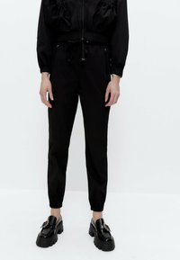 Persoon draagt zwarte joggerbroeken met elastische taille en zwarte chunky instapschoenen met decoratieve gespen, staand tegen een witte achtergrond.