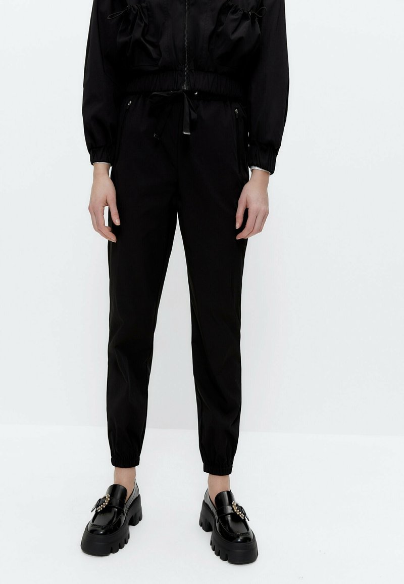 Persoon draagt zwarte joggerbroeken met elastische taille en zwarte chunky instapschoenen met decoratieve gespen, staand tegen een witte achtergrond.