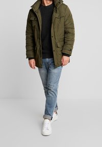 Olivgrön puffjacka med huva, två framfickor och svarta knappar, bärs över en mörk tröja, i kombination med ljusa jeans och vita sneakers.