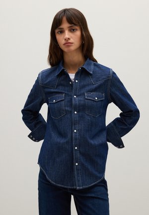 Giovane donna con capelli castani che indossa una camicia di jeans blu scuro con bottoni e jeans abbinati, in piedi con le mani dietro la schiena su uno sfondo neutro.
