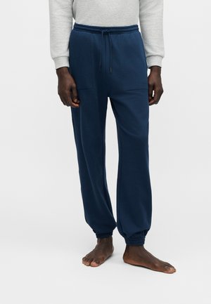 Man draagt marineblauwe joggerbroek en lichtgrijze sweatshirt, staat blootsvoets op een effen witte achtergrond.