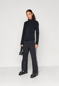 Γυναίκα με μαύρο ζιβάγκο, μαύρο παντελόνι με λευκές ρίγες στο πλάι, μπότες σκι, κρατώντας μαύρα γυαλιά σκι σε απλό φόντο.