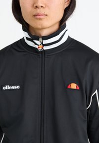 Sort zip-up jakke med stribekrave, hvide detaljer og broderet logo. Materialet ser glat og letvægts ud.