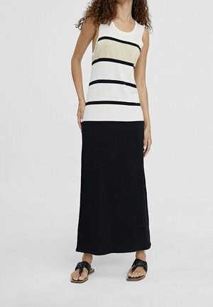 Vrouw in een mouwloos crèmekleurig top met zwarte horizontale strepen en een lange zwarte rok, staand met de handen langs haar zij en zwarte sandalen.