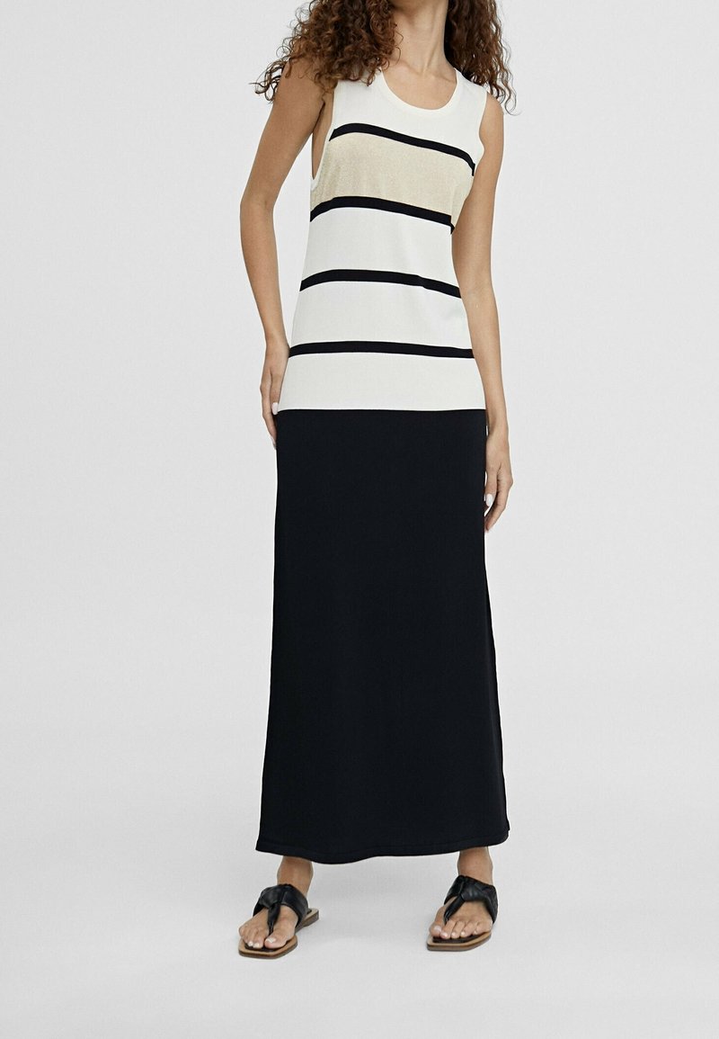 Donna che indossa un top senza maniche color crema con righe orizzontali nere e una lunga gonna nera, in piedi con le mani ai fianchi e sandali neri.