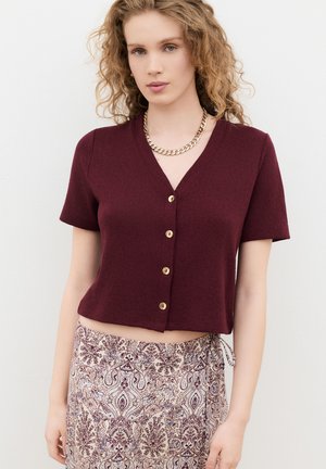 Jonge vrouw met krullend haar, gekleed in een bordeauxrood kort mouw knoopshirt en een beige patroonrok tegen een effen witte achtergrond.
