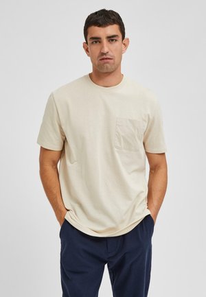 Camiseta de manga corta color crema en tela suave con un bolsillo frontal. Hombros caídos y un corte holgado que aumentan la comodidad.