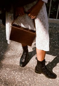 Personne portant une jupe en dentelle blanche et des bottes en cuir noires à lacets, tenant un sac à main en cuir marron foncé, debout sur une surface de gravier.