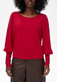 Kobieta w czerwonej bluzce z długim rękawem ozdobionej haftowanymi mankietami oraz ciemnobrązowych skórzanych spodniach, stojąca na tle białym.