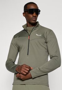 Olivgrüner Pullover mit Viertel-Reißverschluss aus strukturiertem Stoff, schwarzem Reißverschluss und kleinem weißen Logo auf der Brust. Lange Ärmel mit schmalen Bündchen.