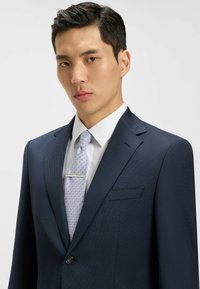 Costume bleu marine à rayures, avec une fermeture à bouton unique, chemise blanche, et cravate à motifs bleu clair avec une pince à cravate en argent.