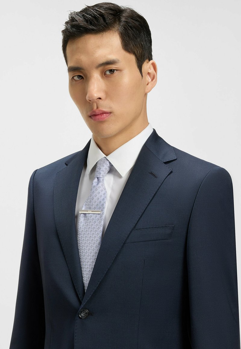 Costume bleu marine à rayures, avec une fermeture à bouton unique, chemise blanche, et cravate à motifs bleu clair avec une pince à cravate en argent.