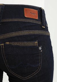 Los jeans de mezclilla azul oscuro cuentan con una etiqueta de cuero marrón con "Pepe Jeans" en la cinturilla trasera y costuras amarillas en contraste en los bolsillos.