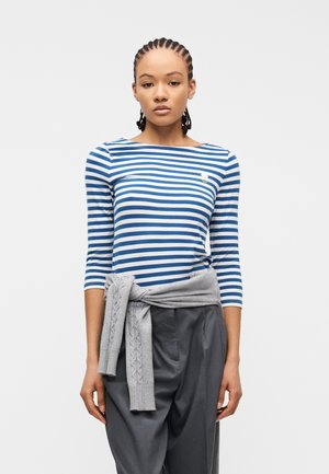 Jeune femme aux cheveux tressés portant un haut rayé bleu et blanc, un pantalon gris et un pull gris clair noué à la taille sur un fond blanc.