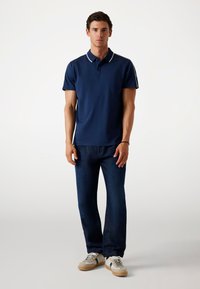 Marinestrick Poloshirt mit weißen Kragenakzenten, kombiniert mit dunkelblauen Denimhosen und beigen Sneakers mit grünen und weißen Details.